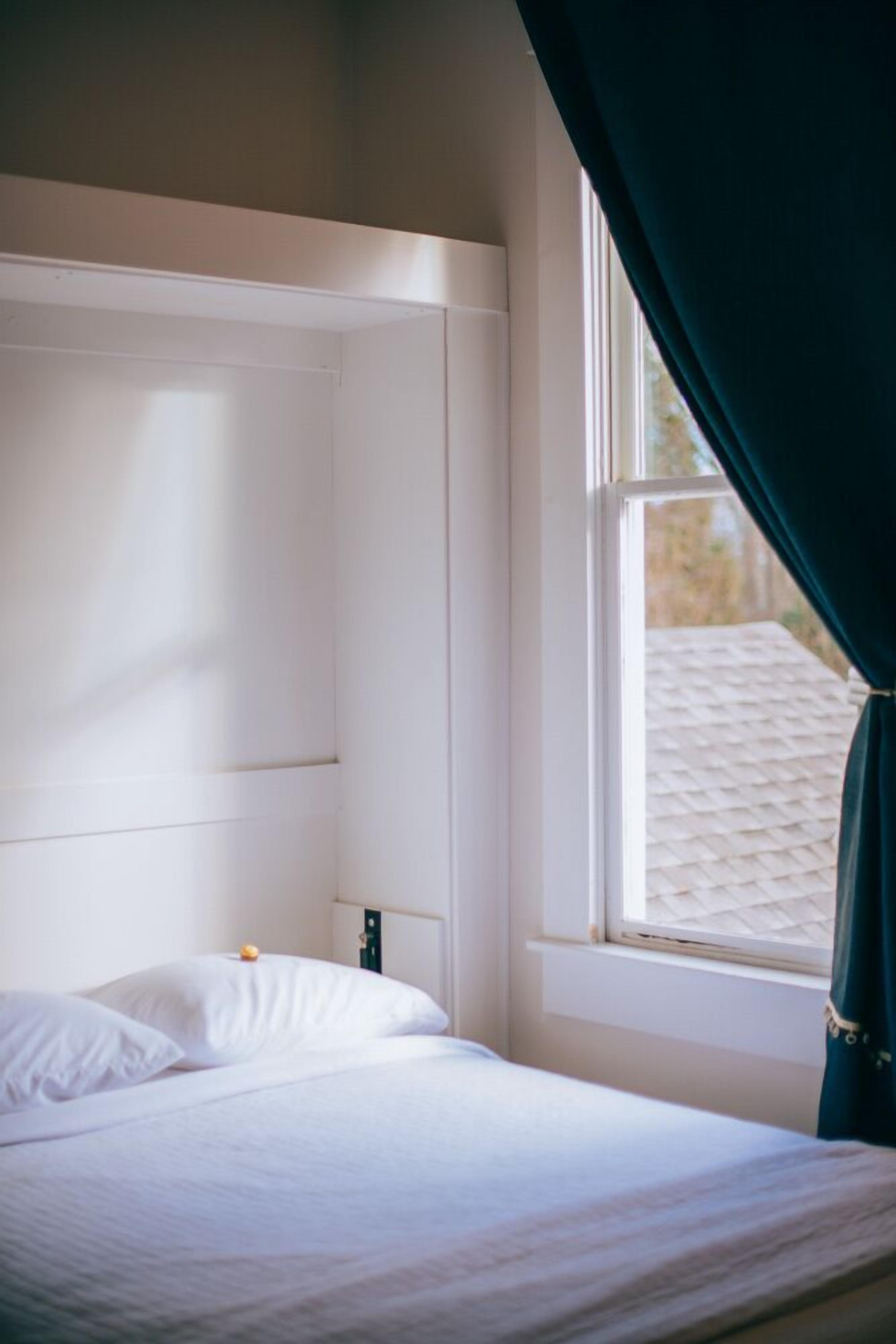 Ein ordentlich bezogenes Bett mit weißen Laken in einem Zimmer mit Fenster und weißem Kopfteil.