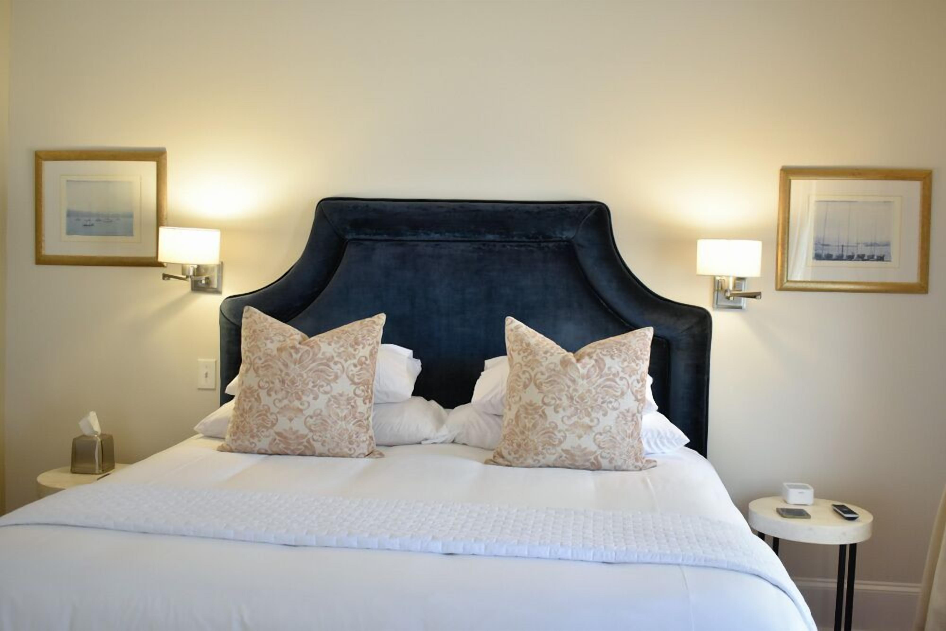 Een keurig opgemaakt bed met een donker hoofdbord, twee decoratieve kussens en een nachtkastje met een lamp en een zakdoos.