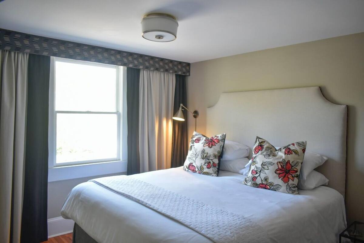 A neatly made bed with floral pillows, a bedside lamp, and a window with curtains.