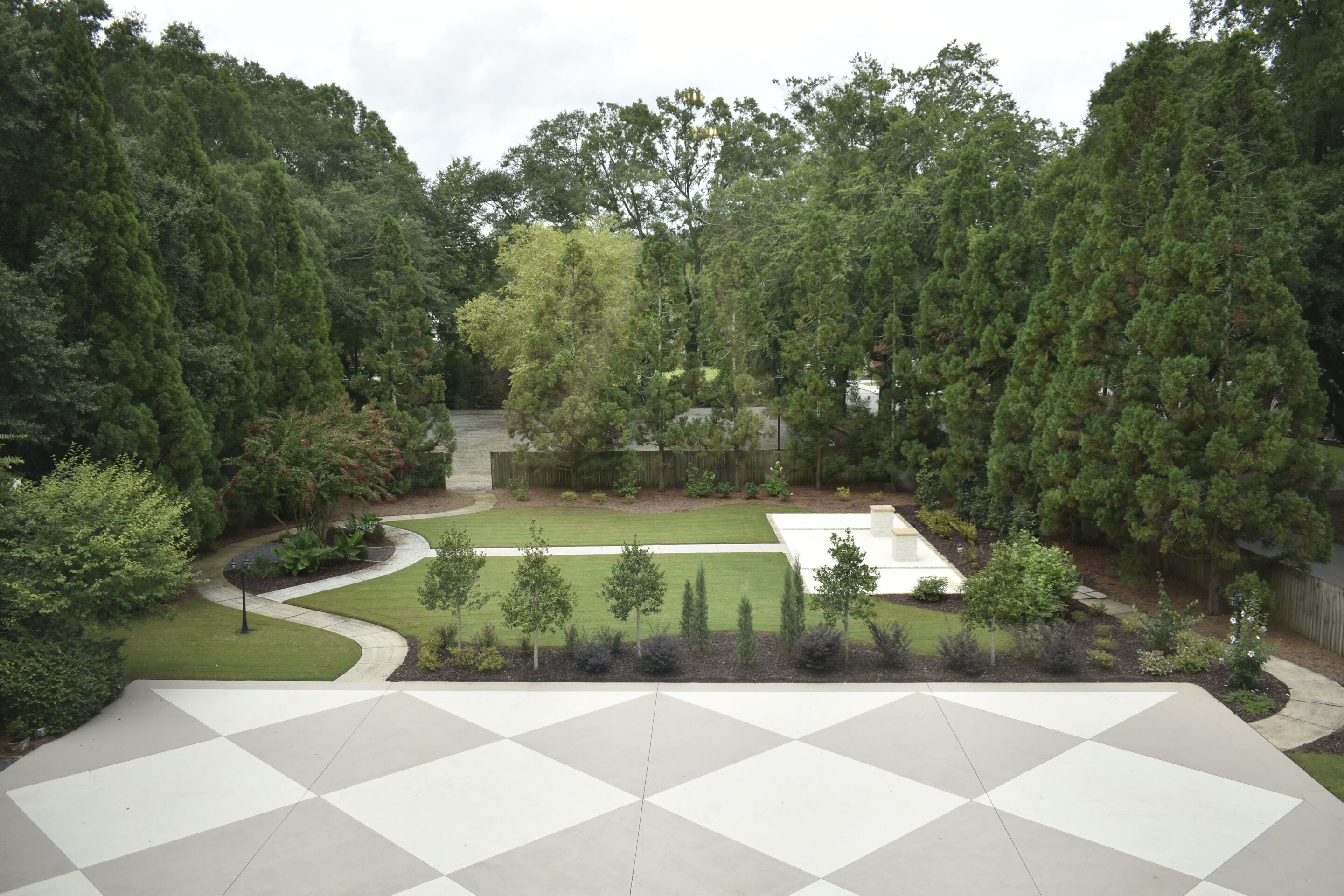 A courtyard with a geometric tile floor, surrounded by dense trees and a small lawn area.