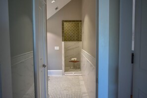 A narrow hallway leading to a bathroom with a tiled floor, a shower area, and a small wooden stool.