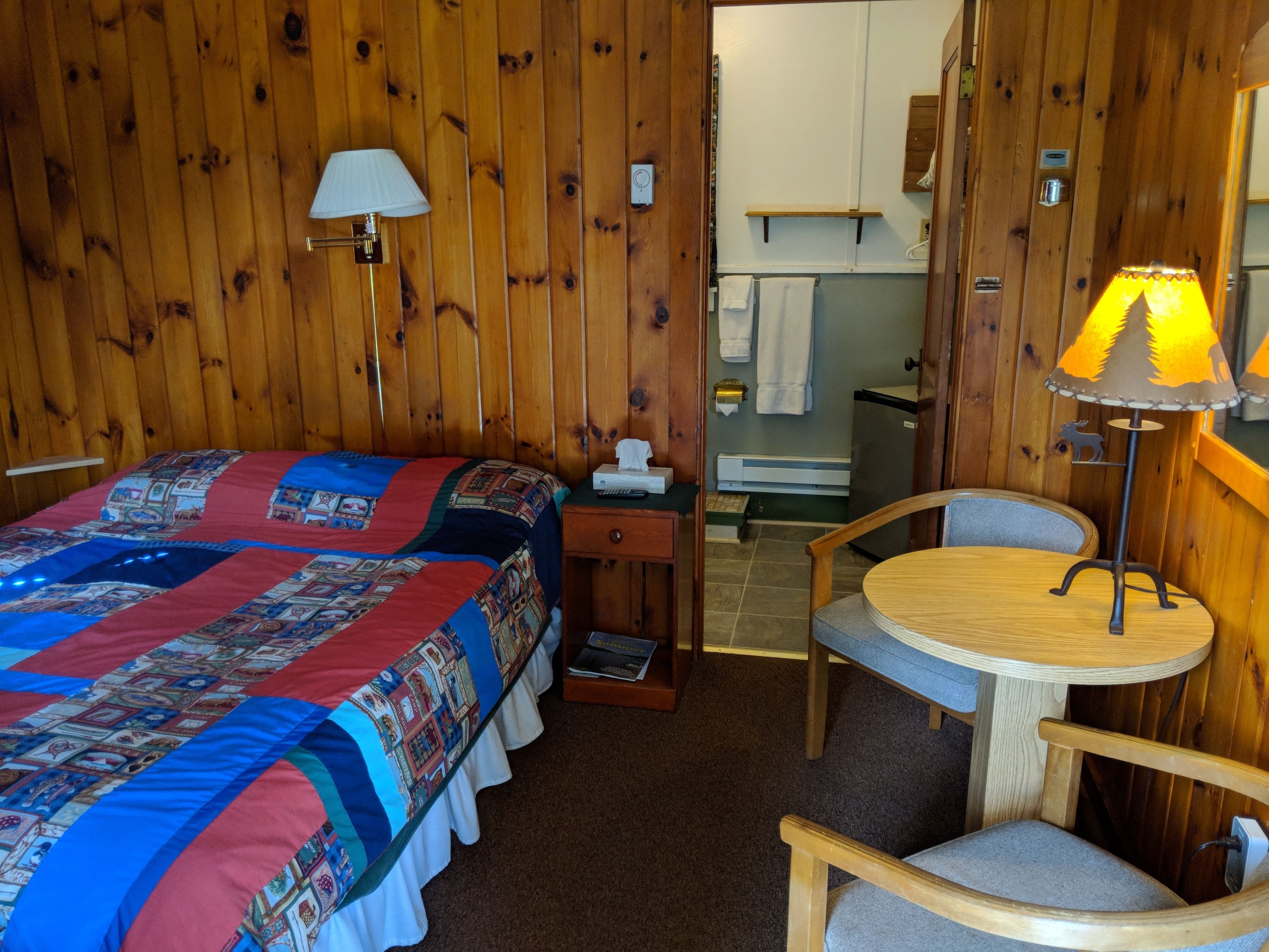 A cozy room with a bed, a chair, a small table, a lamp, and a bathroom visible through an open door.