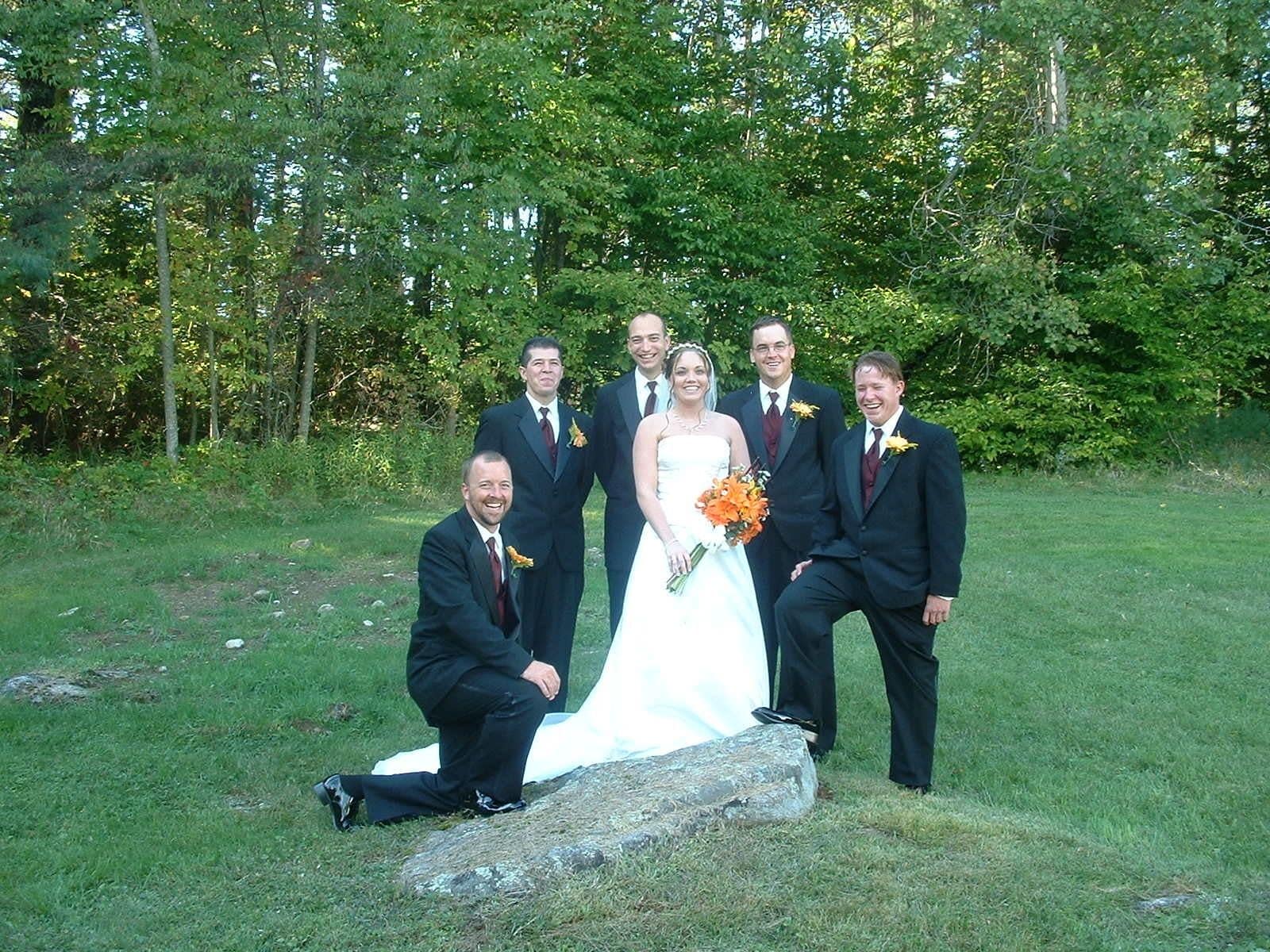 A group of people in formal attire posing outdoors with a grassy area and trees in the background.