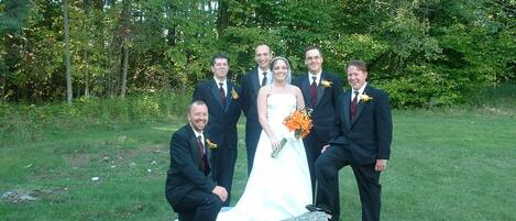 A group of people in formal attire posing outdoors with a grassy area and trees in the background.