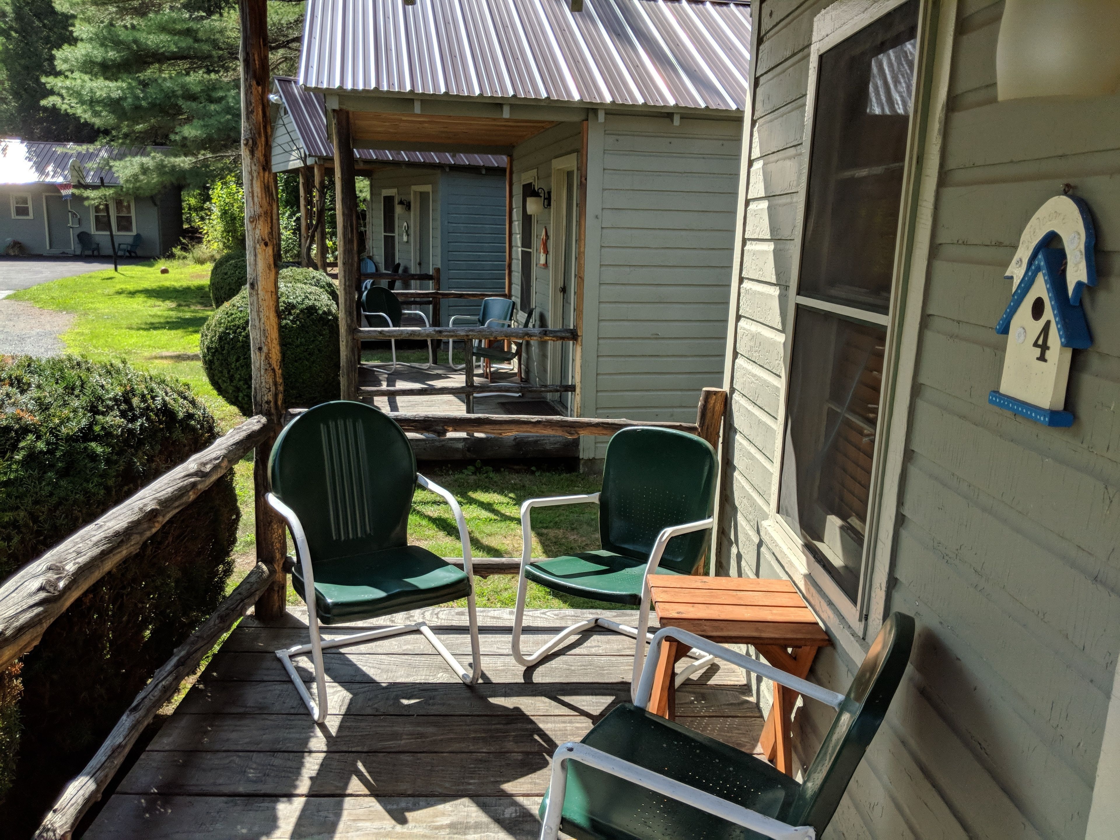 A porch with green chairs and a small table, a wooden deck, and a house with a number 4 birdhouse on the wall.