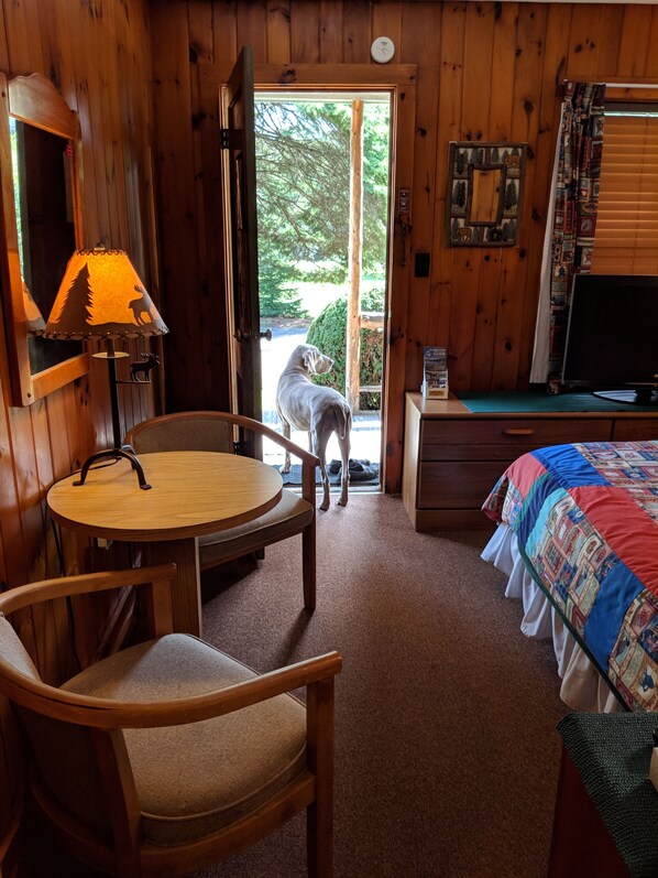 Una habitación de cabaña de madera con una mesa redonda, una silla, una cama con colcha de colores, un televisor y un perro de pie junto a la puerta.