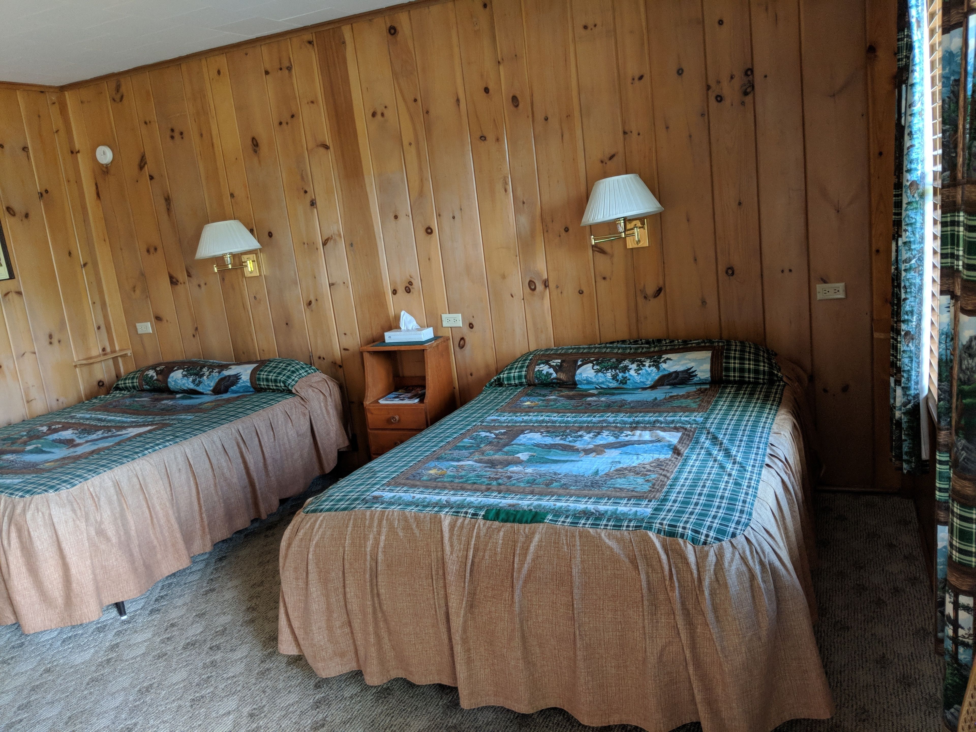 Una habitación con dos camas, paredes de madera y dos lámparas fijadas a la pared.
