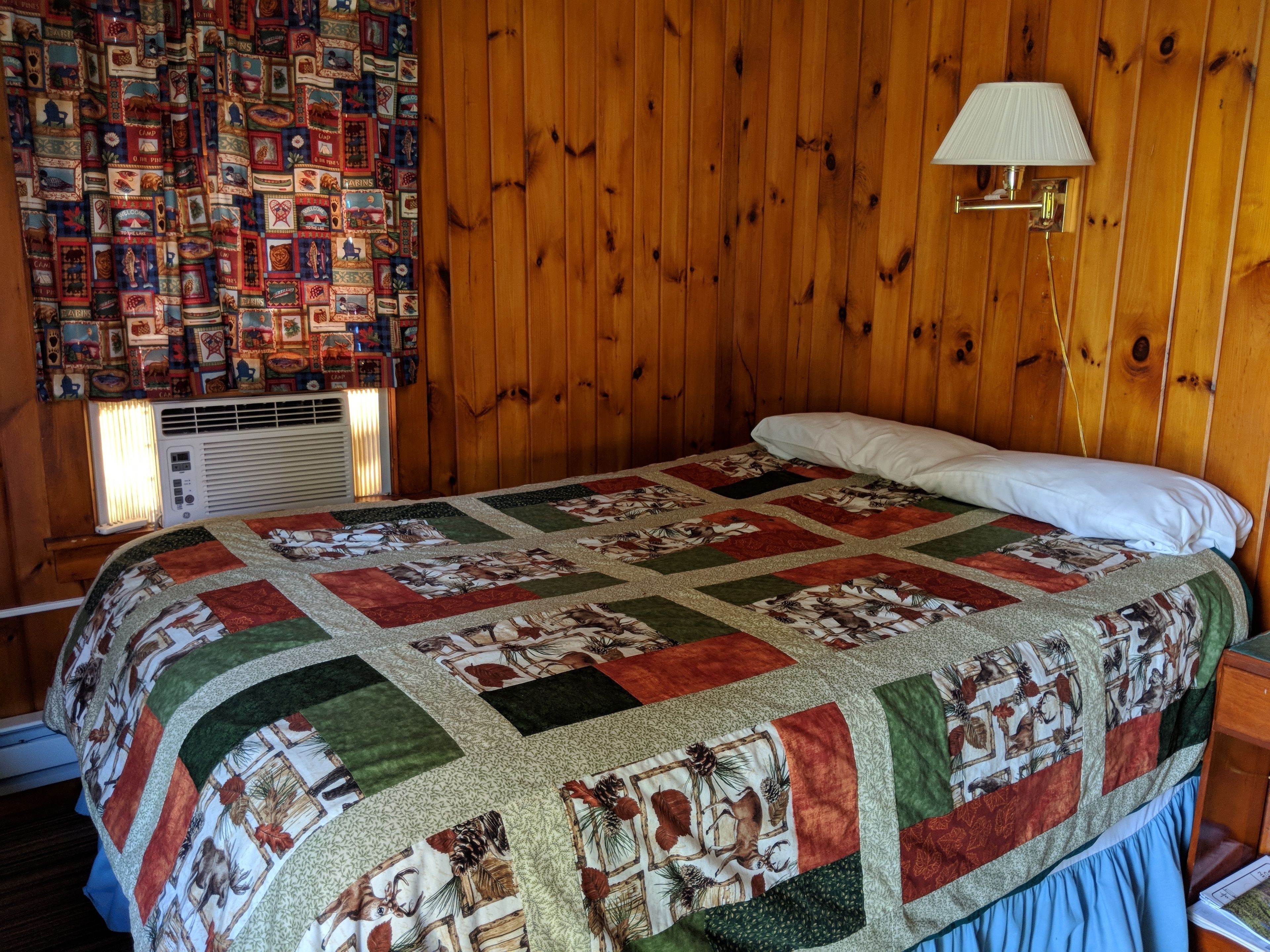 Ein Zimmer mit Holzwänden, einer gesteppten Bettdecke, einer Wandlampe und einem Fenster mit Jalousien.