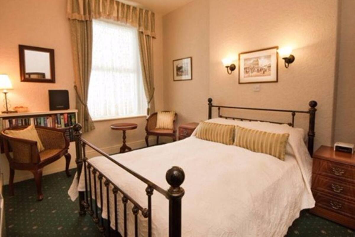 A neatly arranged bedroom with a bed, a chair, a small table, a bookshelf, and a window with curtains.