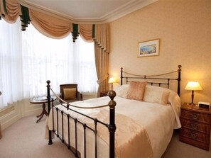 A bedroom with a four-poster bed, a chair, a nightstand, and a window with curtains.