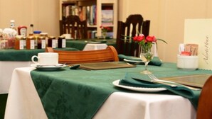 A dining table set with a white tablecloth, a glass of water, a plate, a napkin, a smartphone, and a small vase with red flowers. - Adcote House (Llandudno)