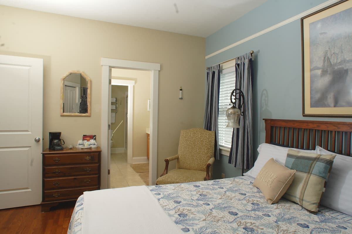 A bedroom with a bed, a dresser, a chair, a window with curtains, and a painting on the wall.