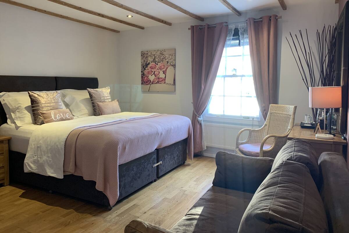 A bedroom with a bed, a nightstand, a chair, a sofa, a window with curtains, and a wall-mounted lamp.