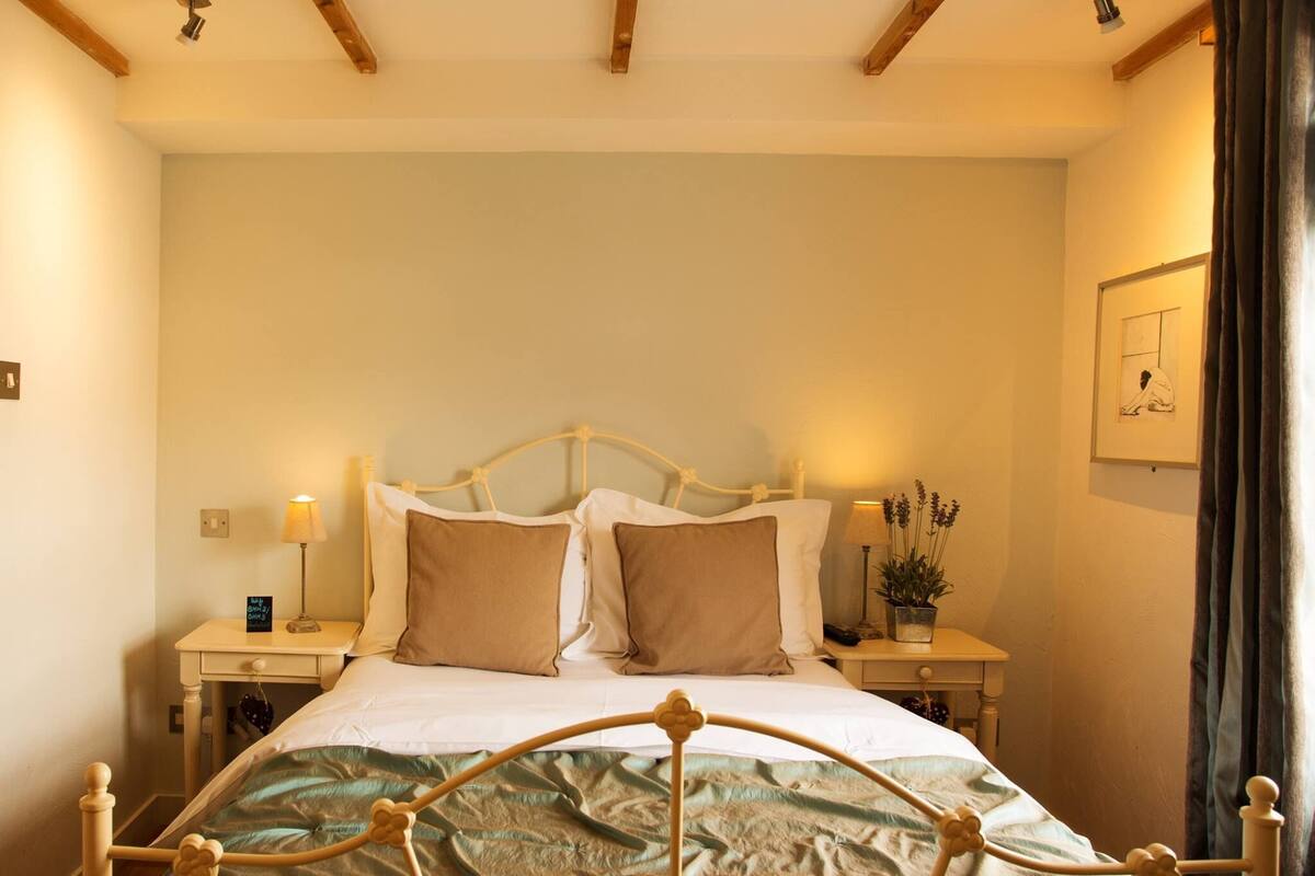 A bedroom with a bed, two bedside tables, a nightstand, a lamp, a potted plant, and a framed picture on the wall.