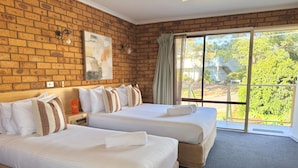 A hotel room with two beds, a brick wall, a painting, and a view of greenery. - Ocean View Motor Inn (Merimbula)