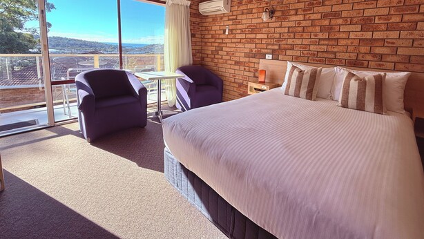 A hotel room with a large bed, two armchairs, a small table, a nightstand, and a view through a glass door.