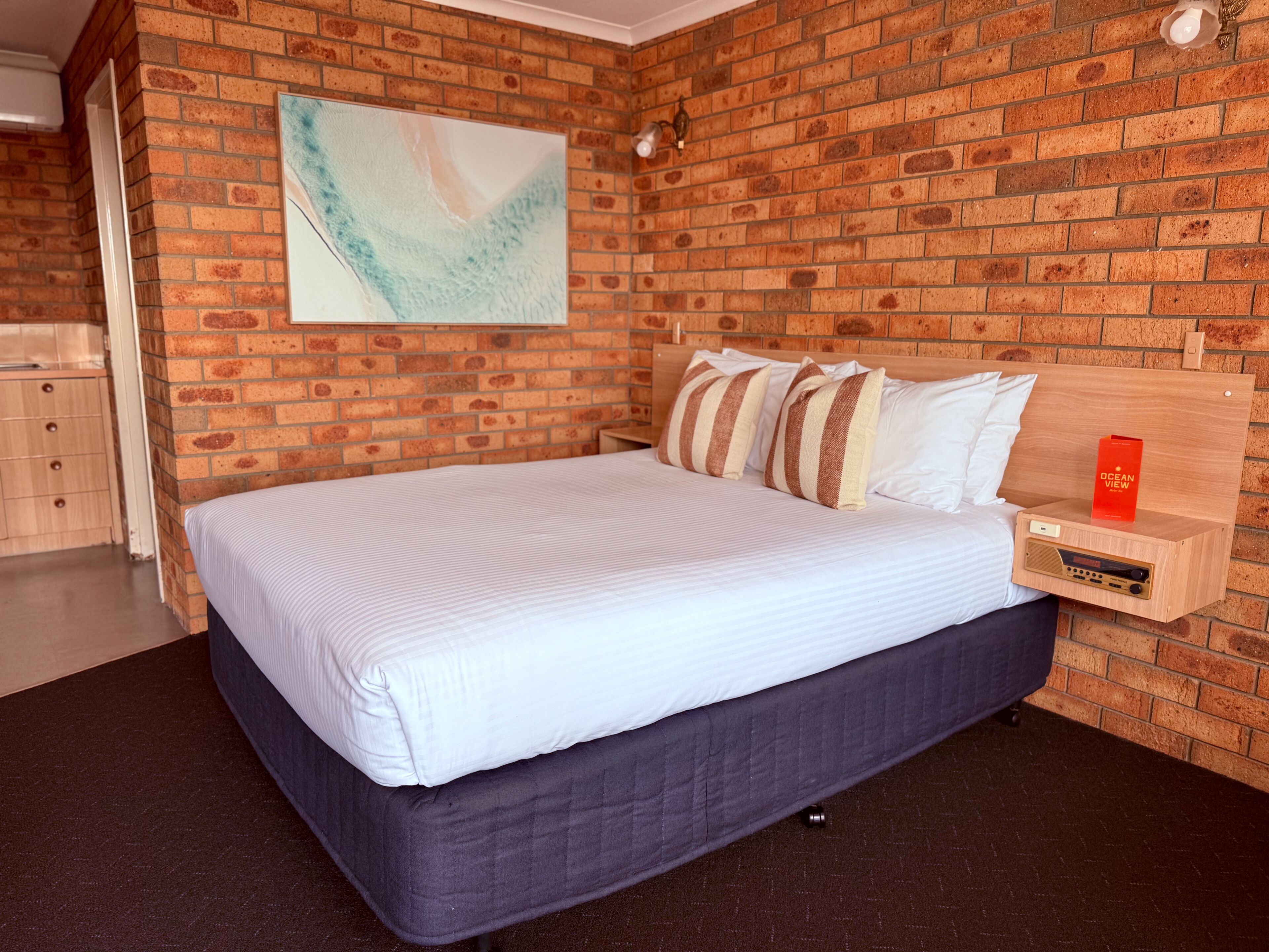 Una habitación con pared de ladrillo, cama con ropa de cama a rayas, mesita de noche con radio y un cuadro en la pared.