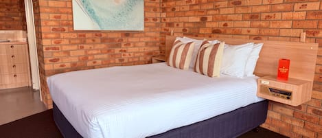 Una habitación con pared de ladrillo, cama con ropa de cama a rayas, mesita de noche con radio y un cuadro en la pared.