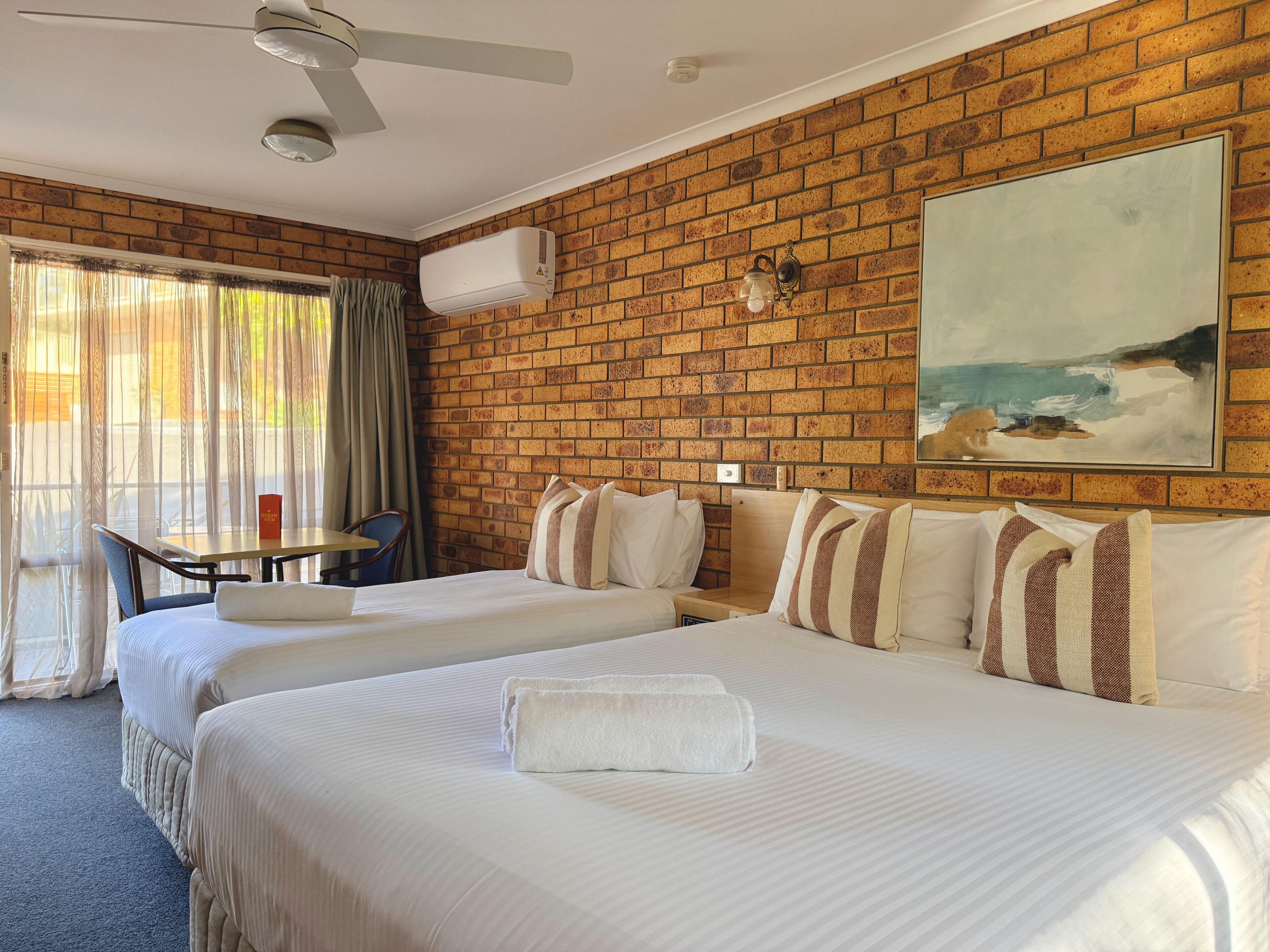 A hotel room with two beds, a table, a chair, a ceiling fan, an air conditioning unit, and a painting on the wall.