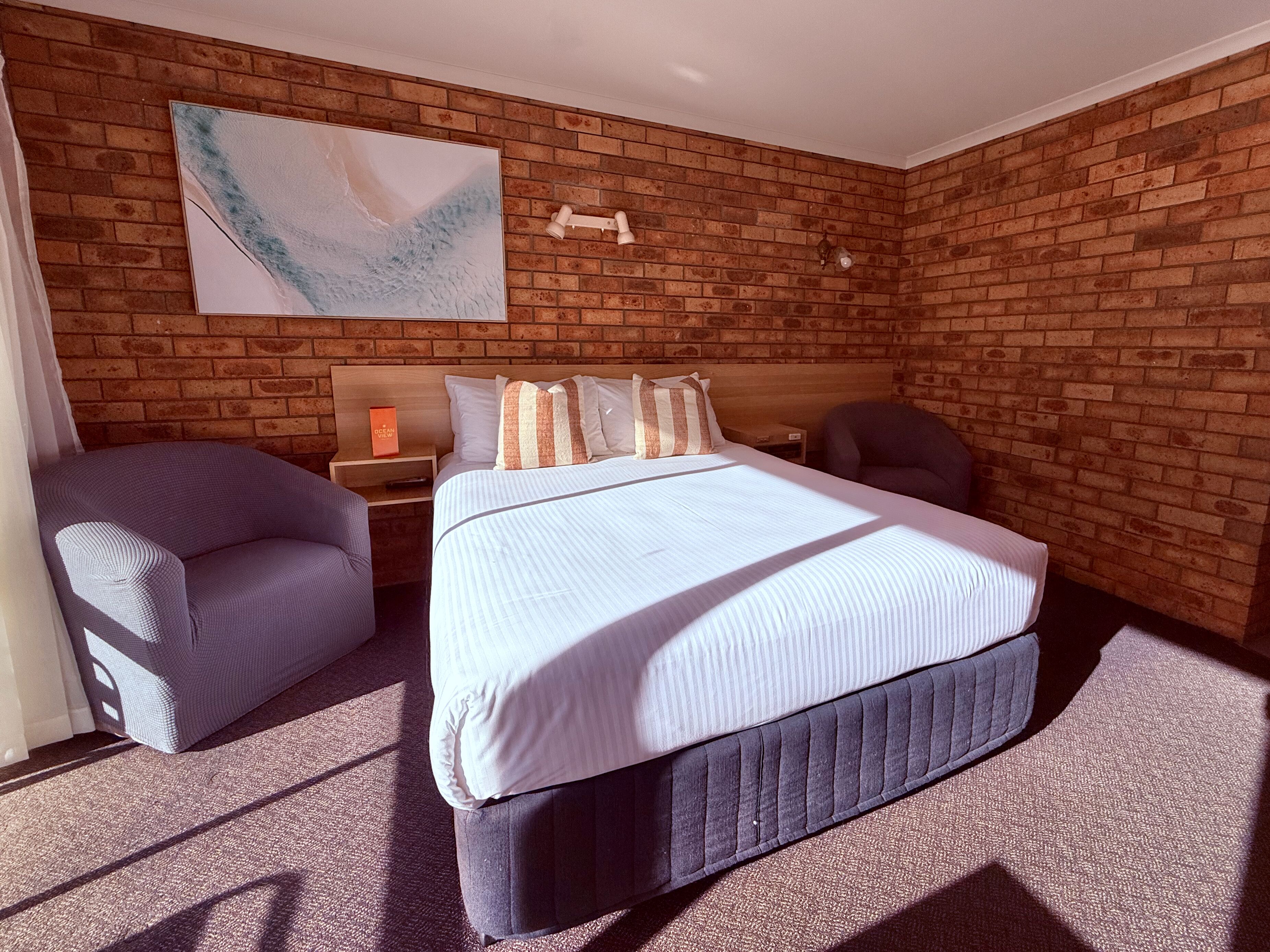 A hotel room with a brick wall, a bed with white bedding, a nightstand, a chair, and a wall-mounted lamp.
