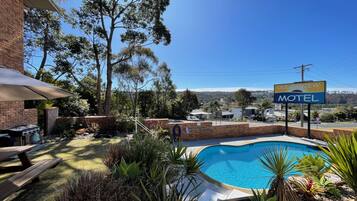A motel with a swimming pool surrounded by plants, a barbecue area, and a view of the landscape.