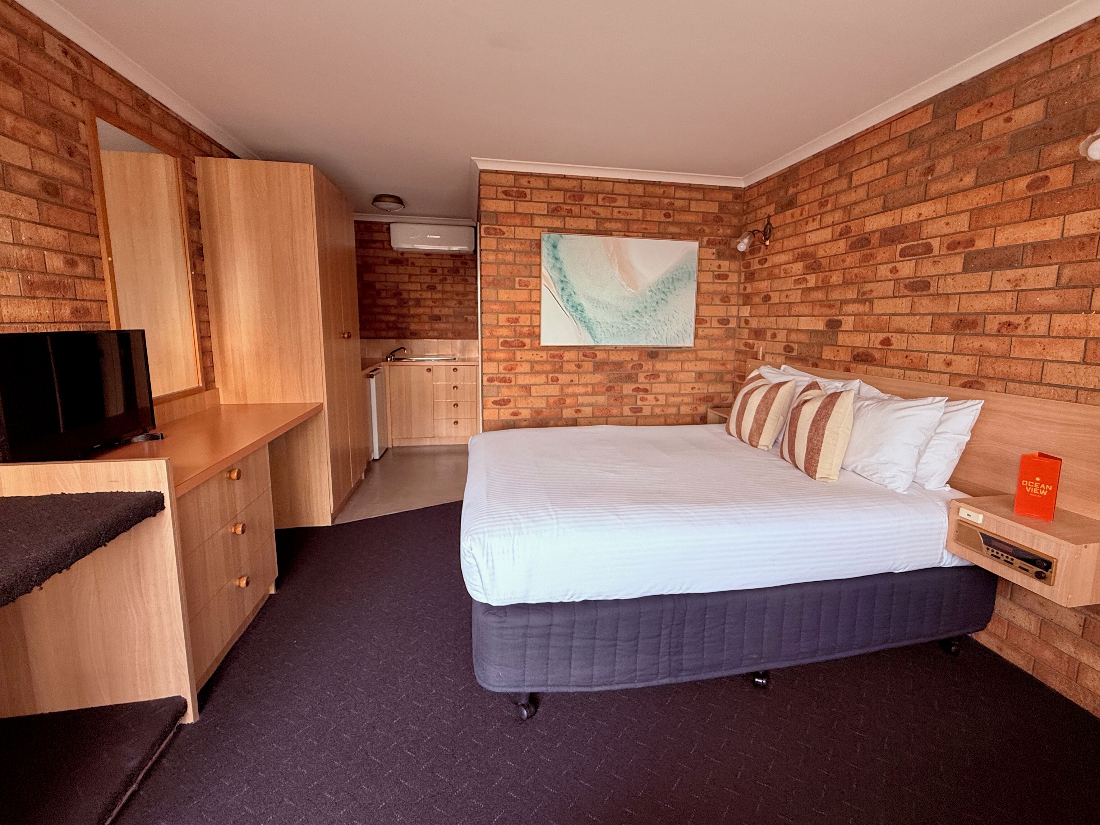 Habitación de hotel con pared de ladrillo, cama con ropa blanca, escritorio de madera con televisor y una pequeña kitchenette.