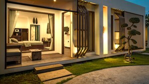A modern house with a well-lit interior, a visible bedroom with a bed and seating area, and a small outdoor garden with a tree. - Villa Nero (Gili Trawangan)