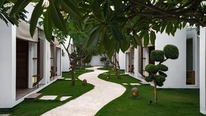 A pathway leading through a well-maintained garden with neatly trimmed hedges and a white building with multiple doors. - Villa Nero (Gili Trawangan)