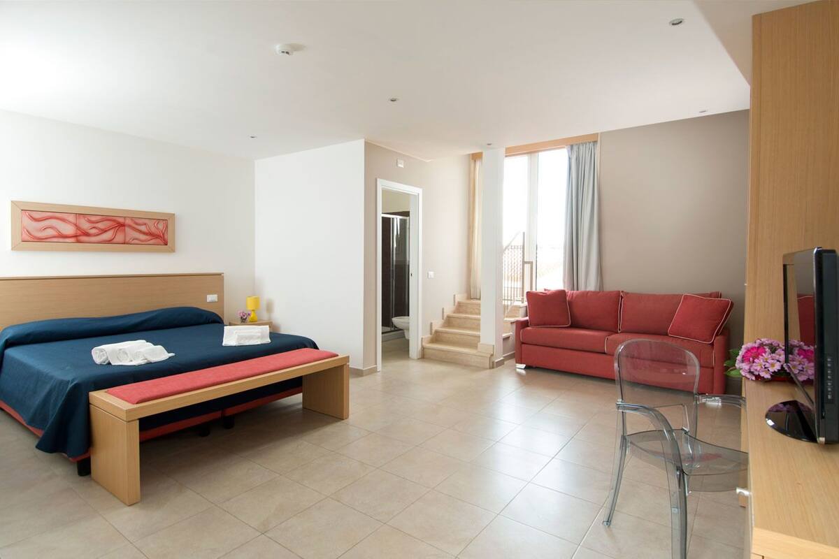 A modern hotel room with a bed, a red sofa, a TV, and a bathroom visible through an open door.