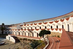 A building with a curved roof and multiple arches, surrounded by a sandy area with some vegetation.