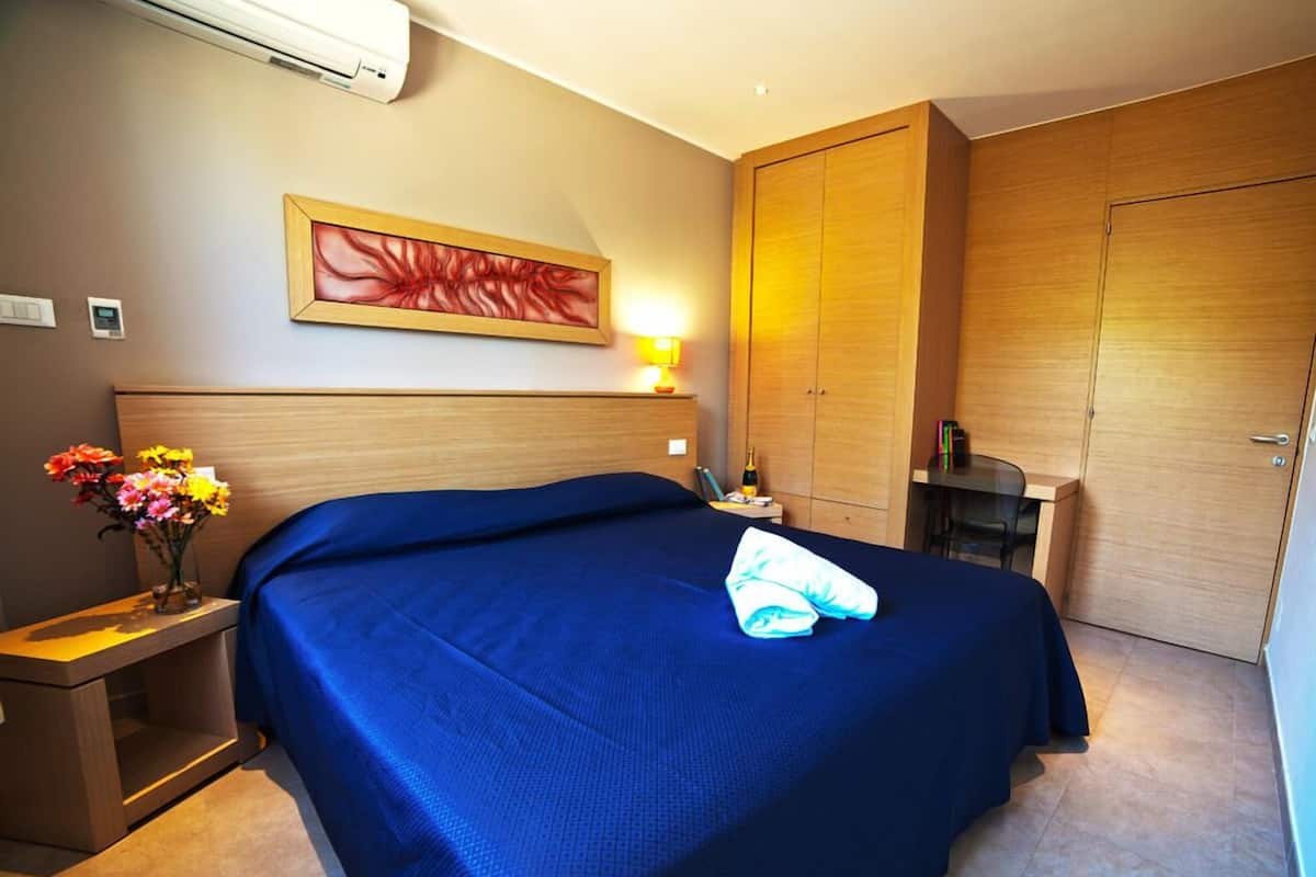 A hotel room with a large bed, a wooden wardrobe, a desk with a lamp, and a painting on the wall.