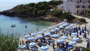Una platja amb nombrosos para-sols i gandules, una costa rocosa i un edifici amb arcs al fons.