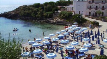 Una playa con numerosas sombrillas y tumbonas, un litoral rocoso y un edificio con arcos al fondo.