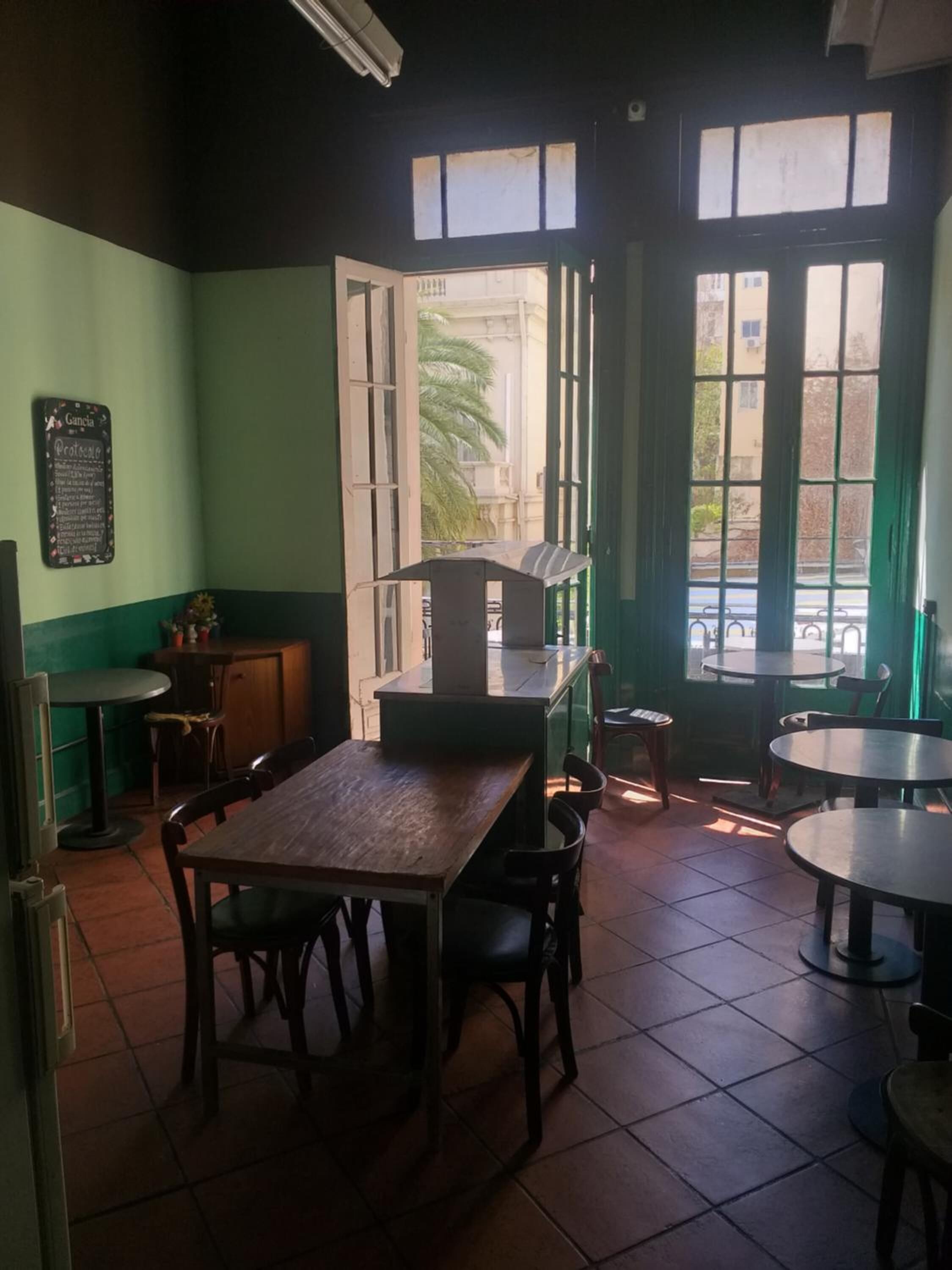 L’intérieur d’un restaurant avec une table et des chaises en bois, un tableau noir servant de menu, et de grandes fenêtres donnant sur la rue.