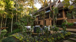 A two-story building with a brick exterior, surrounded by lush greenery and a variety of flowering plants. - Vanā Som (Mae Rim)