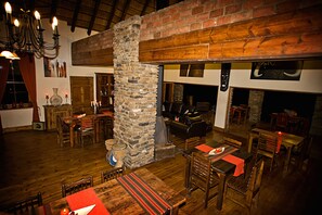 A rustic interior with wooden flooring, stone pillars, and a brick fireplace. Tables and chairs are arranged for dining. - Addo Bush Palace Private Reserve (Addo)