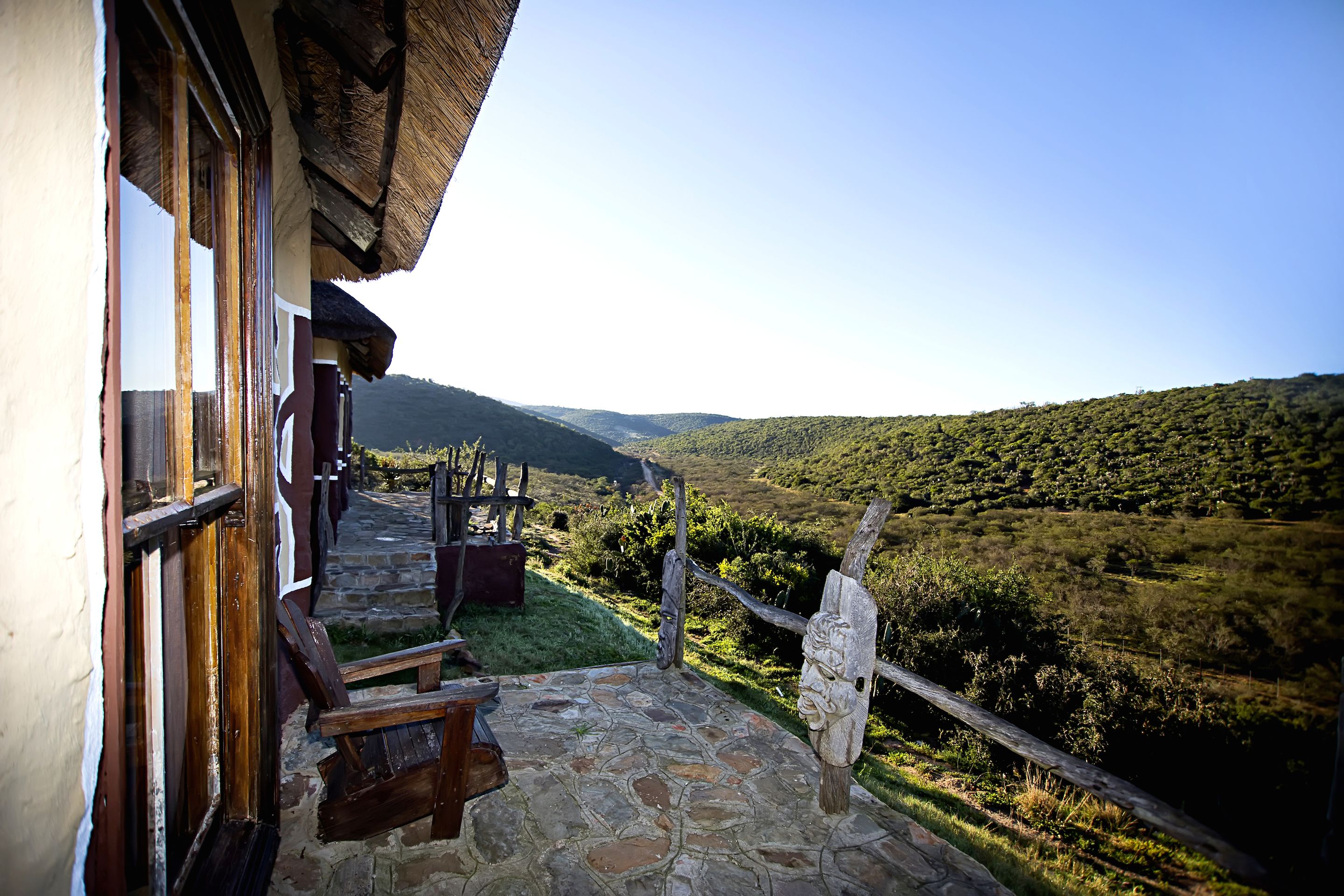 A stone-paved porch with wooden benches, a rustic wooden fence, and a view of a lush valley.