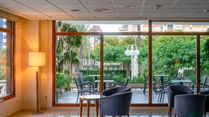Un espacio interior moderno con zona de estar, ventanas amplias, una lámpara de pie y vistas a un patio exterior con mesas y sillas.