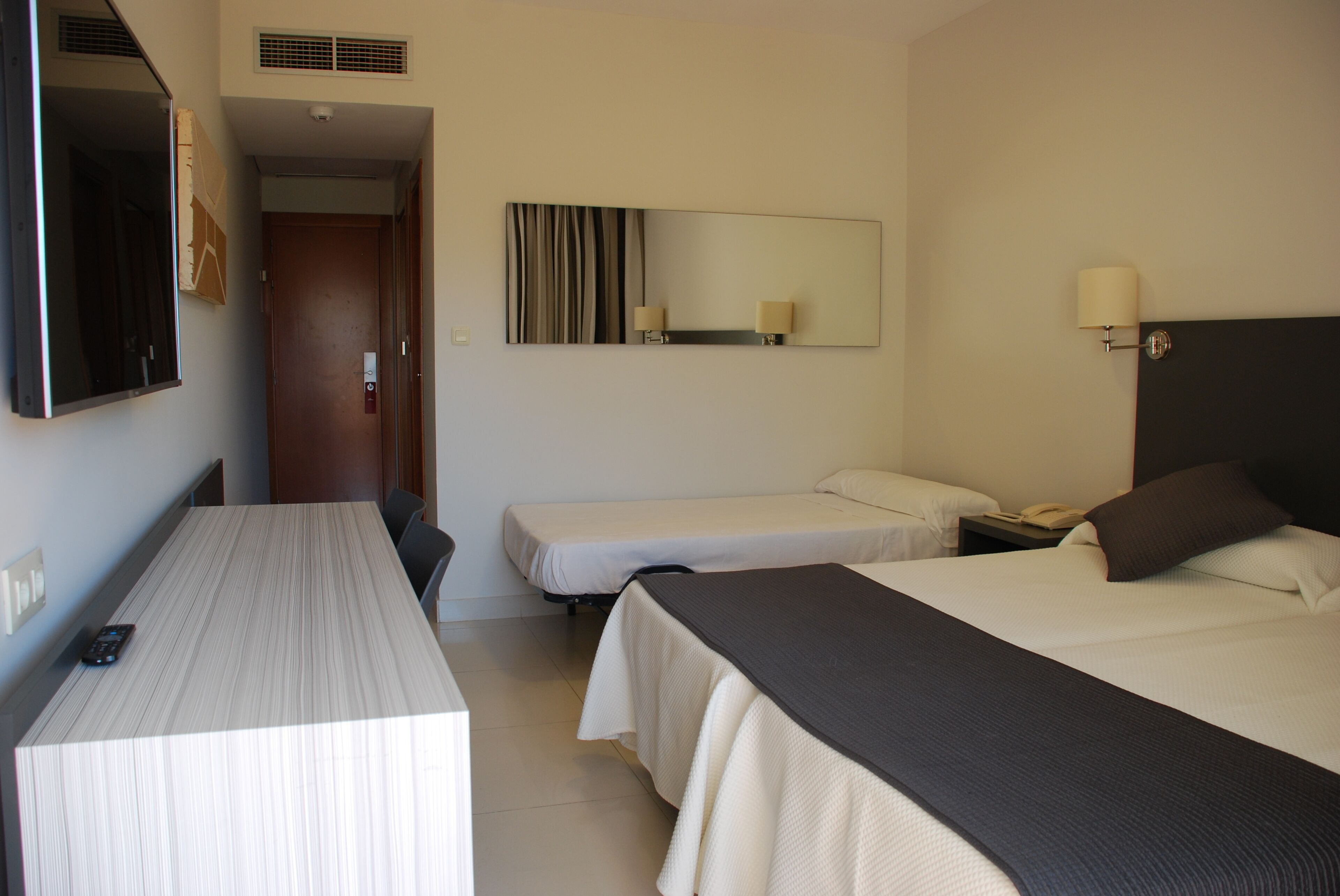 A hotel room with a bed, a desk, a chair, a mirror, and a bedside table with a lamp.