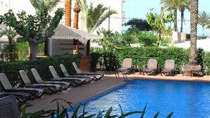 Un área junto a la piscina con sillones, una estructura de techo de paja y palmeras.