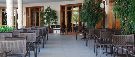 A covered outdoor seating area with wicker chairs and tables, large windows, and potted plants.
