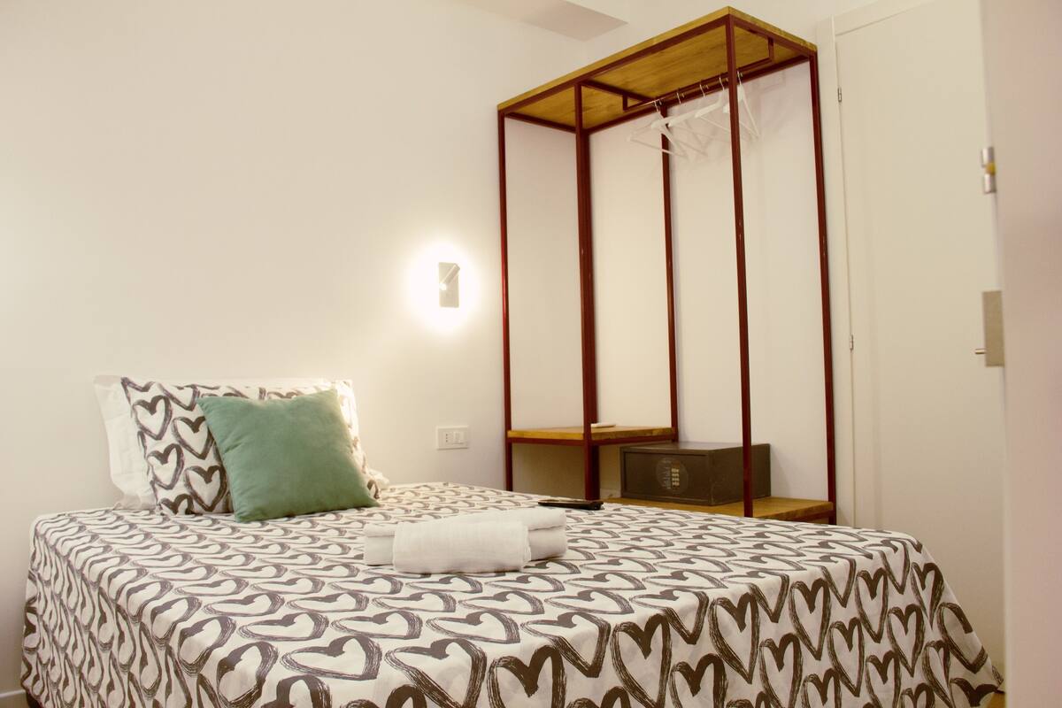 A neatly made bed with patterned bedding, a bedside table, a wall-mounted light, and a wooden canopy structure.