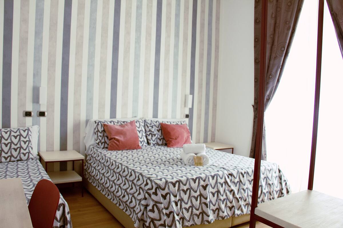 A hotel room with a striped wall, a bed with patterned bedding, a small table, and a chair.