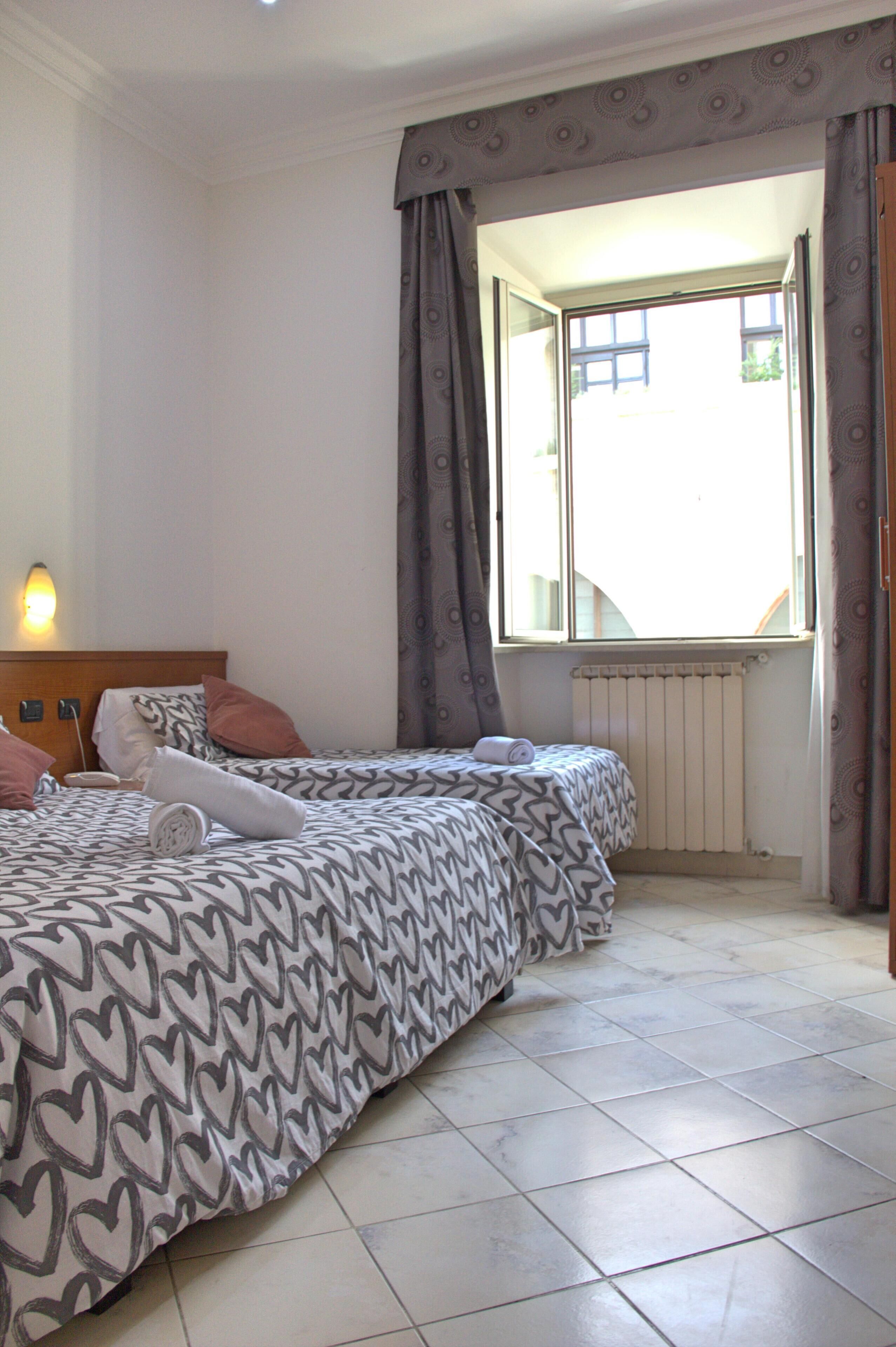 A hotel room with two beds, a window with curtains, a radiator, and a wall-mounted light.
