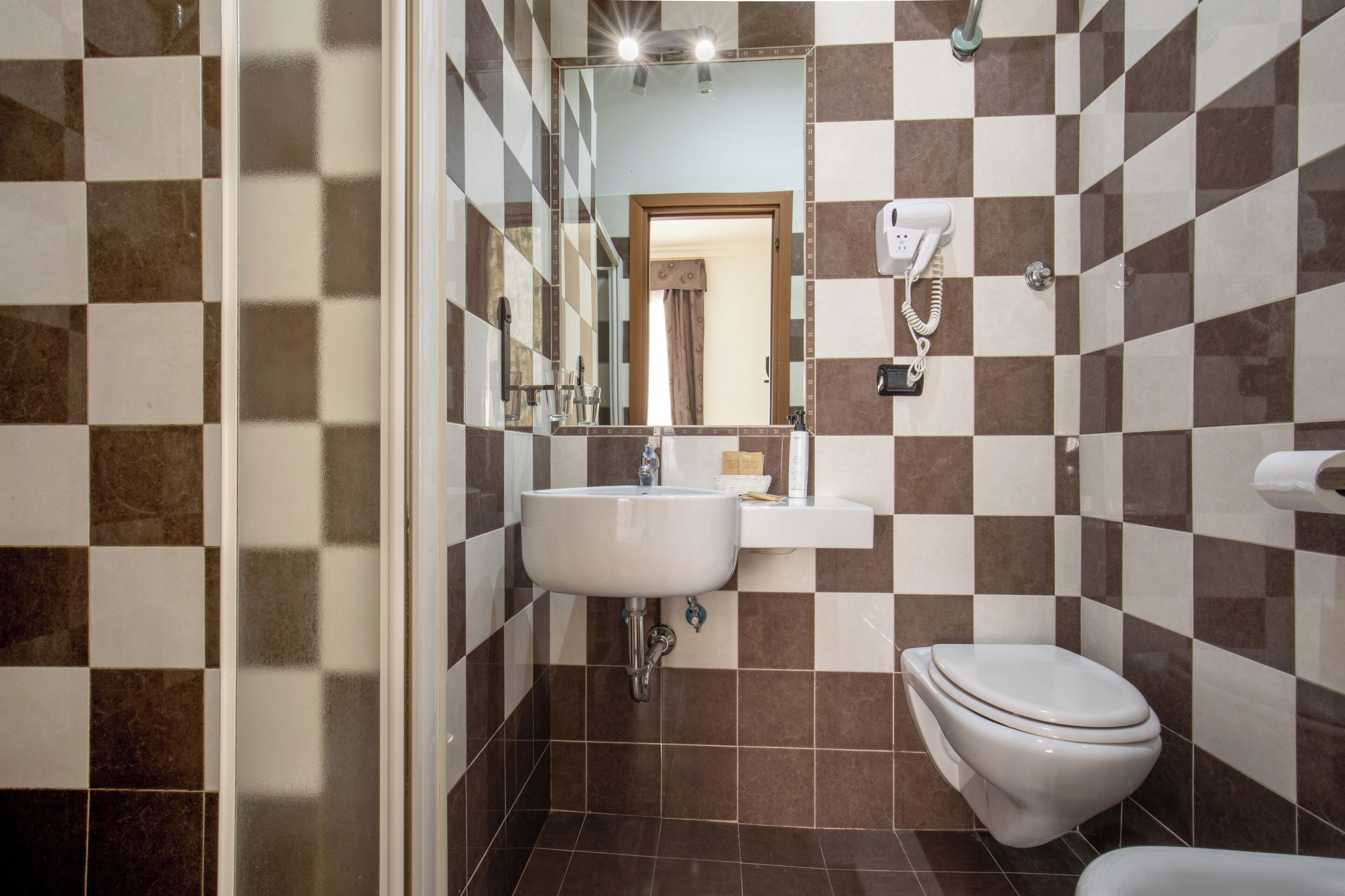 A bathroom with a checkered tile pattern, a sink, a toilet, and a hairdryer mounted on the wall.