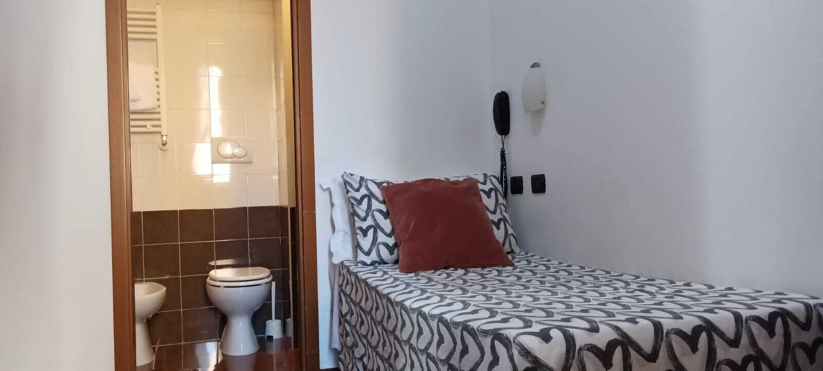 A small room with a bed, a red pillow, a white wall, and a toilet.