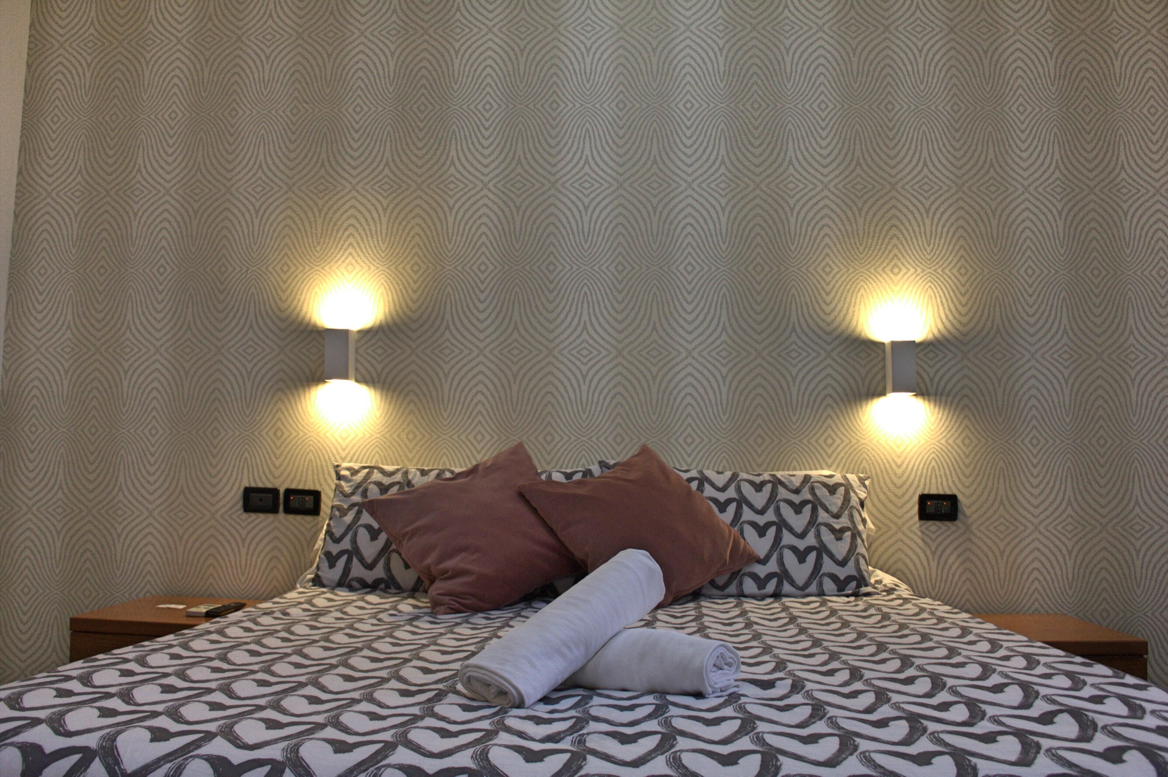 A neatly made bed with patterned bedding, two wall-mounted lamps, and a patterned wall.