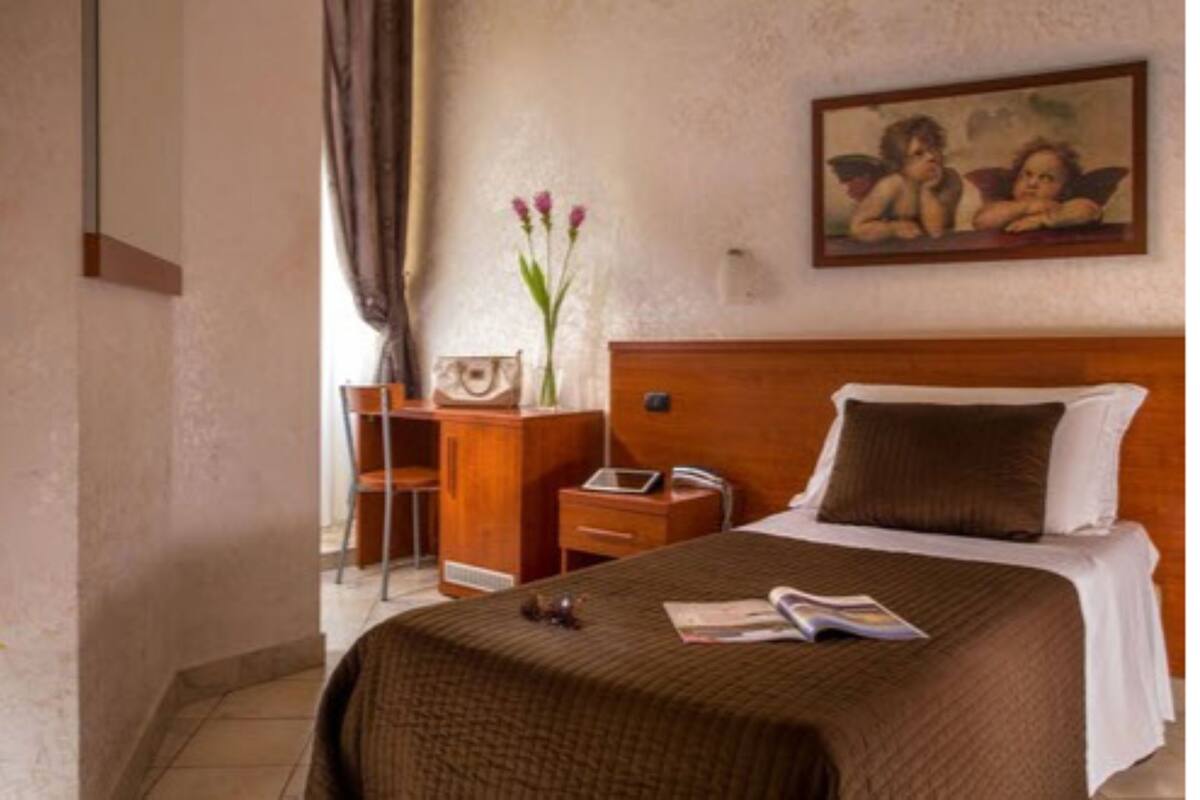 A hotel room with a large bed, a wooden nightstand, a desk with a chair, a vase with flowers, and a painting on the wall.
