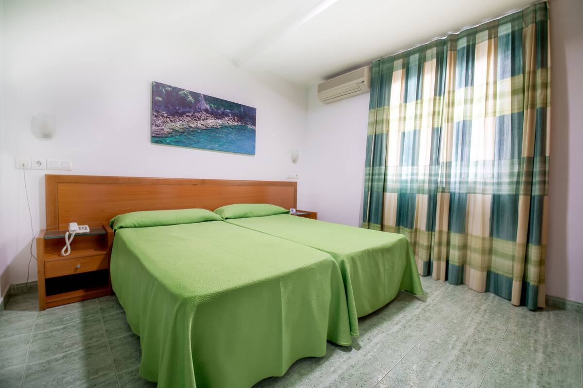 A room with a bed, a bedside table with a phone, a painting on the wall, and a window with curtains.