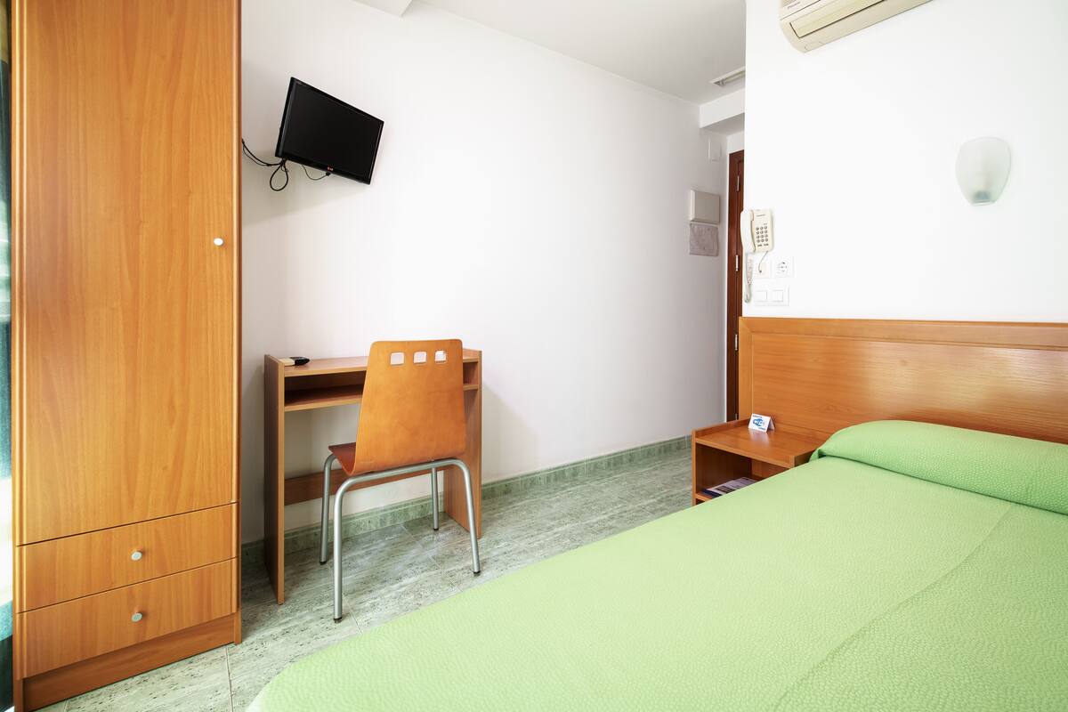 A hotel room with a bed, a desk, a chair, a wardrobe, a television, a bedside table, and a nightstand.